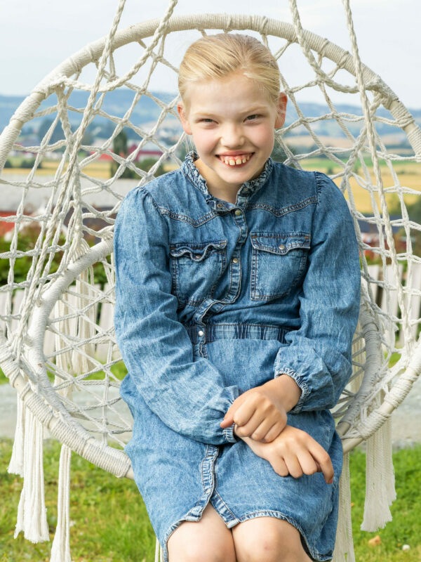 Carmen sitter i en kurvstol med blå denimkjole. Hun smiler til fotografen