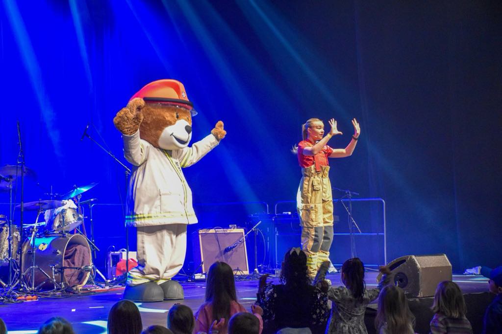 Brannbamsen Bjørnis og en dame står og danser med hendene over hodet på en scene, med barn fremfor seg. Bakgrunnen er blå.