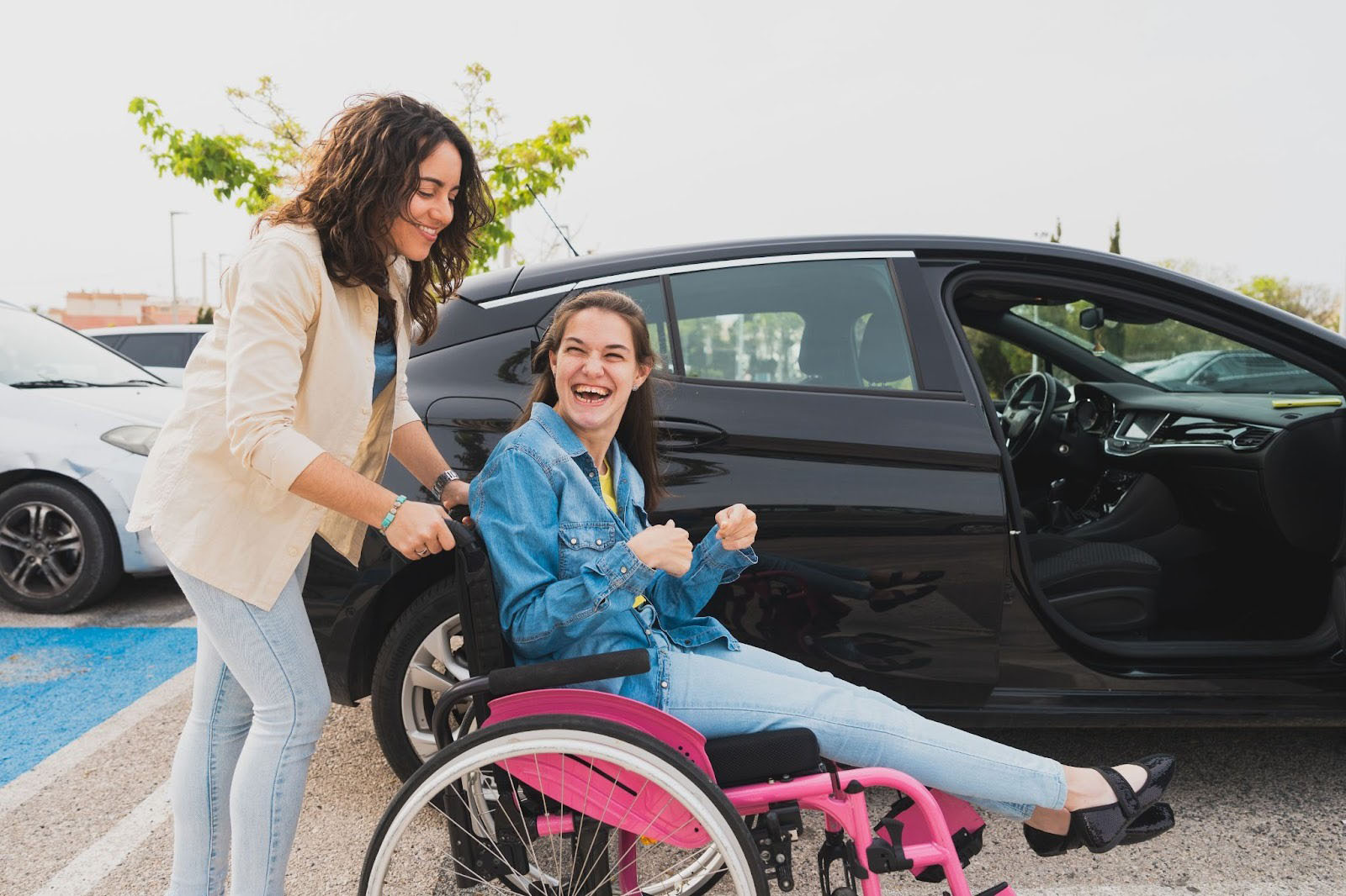 En BPA-assistent står bak en rosa rullestol og hjelper en ung kvinne som sitter i rullestol ved en åpen bil i en parkeringsplass.