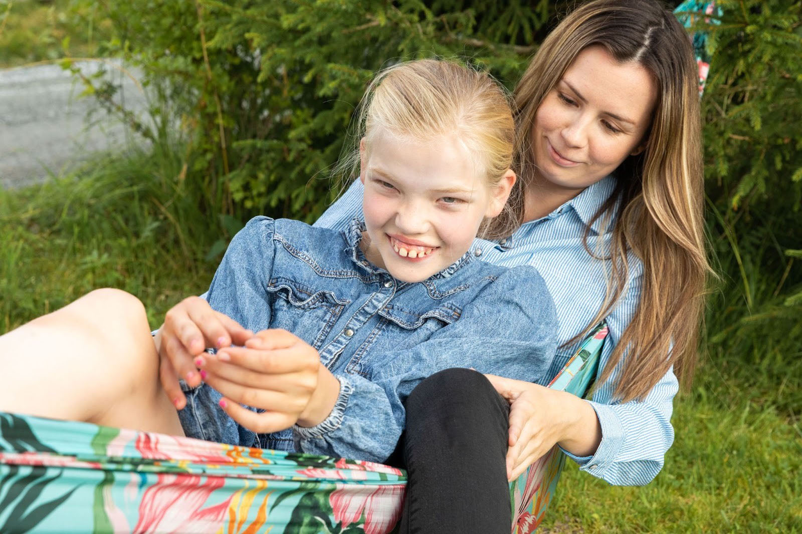 En jente sitter avslappet i fanget til en BPA-assistent ute i hagen, og smiler mot kameraet.